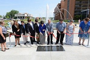 Thomas A. Bruce, M.D., and family members participate in a ribbon-cutting ceremony to dedicate a fountain donated in memory of his wife, Dolores. They stand in a line, holding a red ribbon, with some holding scissors, ready to cut. The fountain, with water spraying upward, is in the background, and a plaque reading "In Loving Memory of Dolores Bruce" is embedded in the ground. The event takes place outdoors on the UAMS campus, with a brick building and a crowd of attendees visible under a clear blue sky.
