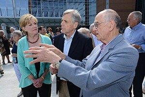 Thomas A. Bruce, M.D., on the right in a light blue suit, engages in conversation with Billie and Skip Rutherford about the newly dedicated fountain in memory of Dolores Bruce. They stand outdoors on the UAMS campus, with a modern glass and brick building in the background. A crowd of attendees is gathered, some walking and others observing the event, under a clear blue sky, creating a lively and commemorative atmosphere.