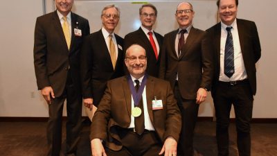 David Ussery, Ph.D. sits in the chair given to him as part of the investiture ceremony. Behind him are UAMS Chancellor Dan Rahn, M.D., ARA President and CEO Jerry Adams, Fred Prior, Ph.D., UAMS Executive Vice Chancellor and Dean of the College of Medicine Pope L. Moseley, M.D., and Søren Brunak, Prof. Ph.D.