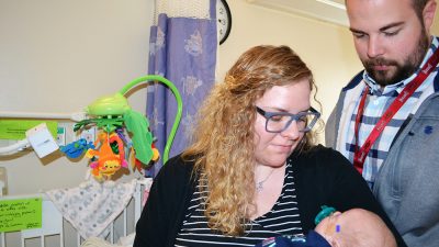 Chelsea Grizzelle, left, and Aaron Grizzelle, right, cradle and visit with their daughter, Gemma, in the UAMS NICU. Gemma's crib and the Angel Eye camera recently installed on it are visible to the left.