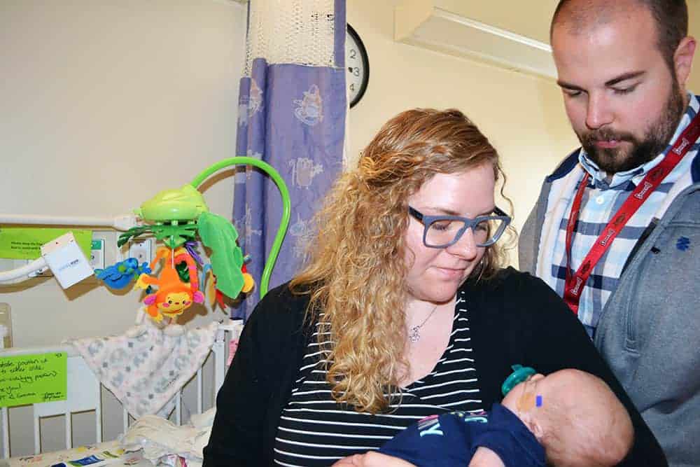 Chelsea Grizzelle, left, and Aaron Grizzelle, right, cradle and visit with their daughter, Gemma, in the UAMS NICU. Gemma's crib and the Angel Eye camera recently installed on it are visible to the left.