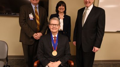 James McDonald, M.D. sits in the chair given to him as part of the investiture, shown here with (from left) Richard Turnage, M.D., UAMS Interim Chancellor Stephanie Gardner, Pharm.D., Ed.D., and Christopher Westfall, M.D.