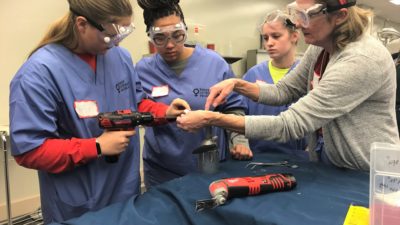 Laurie Hughes, M.D., (right) a retired orthopaedic surgeon, assists female high school students as they make repairs to simulated bones and joints.