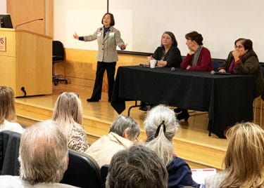 Jeanne Wei, standing on stage, moderates a panel discussion with, left to right, Lou Ann Eads, Denise Compton and Gohit Azhar.
