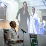Professor in foreground while student dons white coat on screen behind