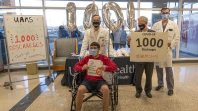 Michael Kent, seated left, holds a sign announcing he is the 1,000th COVID-19 patient to be discharged by the UAMS Medical Center. Behind Kent, left to right, are UAMS Chancellor Cam Patterson, UAMS Medical Center CEO Steppe Mette and hospitalist Franklin Gray.