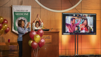 Madison Morris celebrates with family onscreen while Sharanda Williams and Sara Tariq, M.D., watch from the podium in the auditorium.