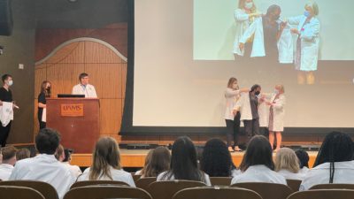 Students walk across the stage one by one to be helped into their white coats