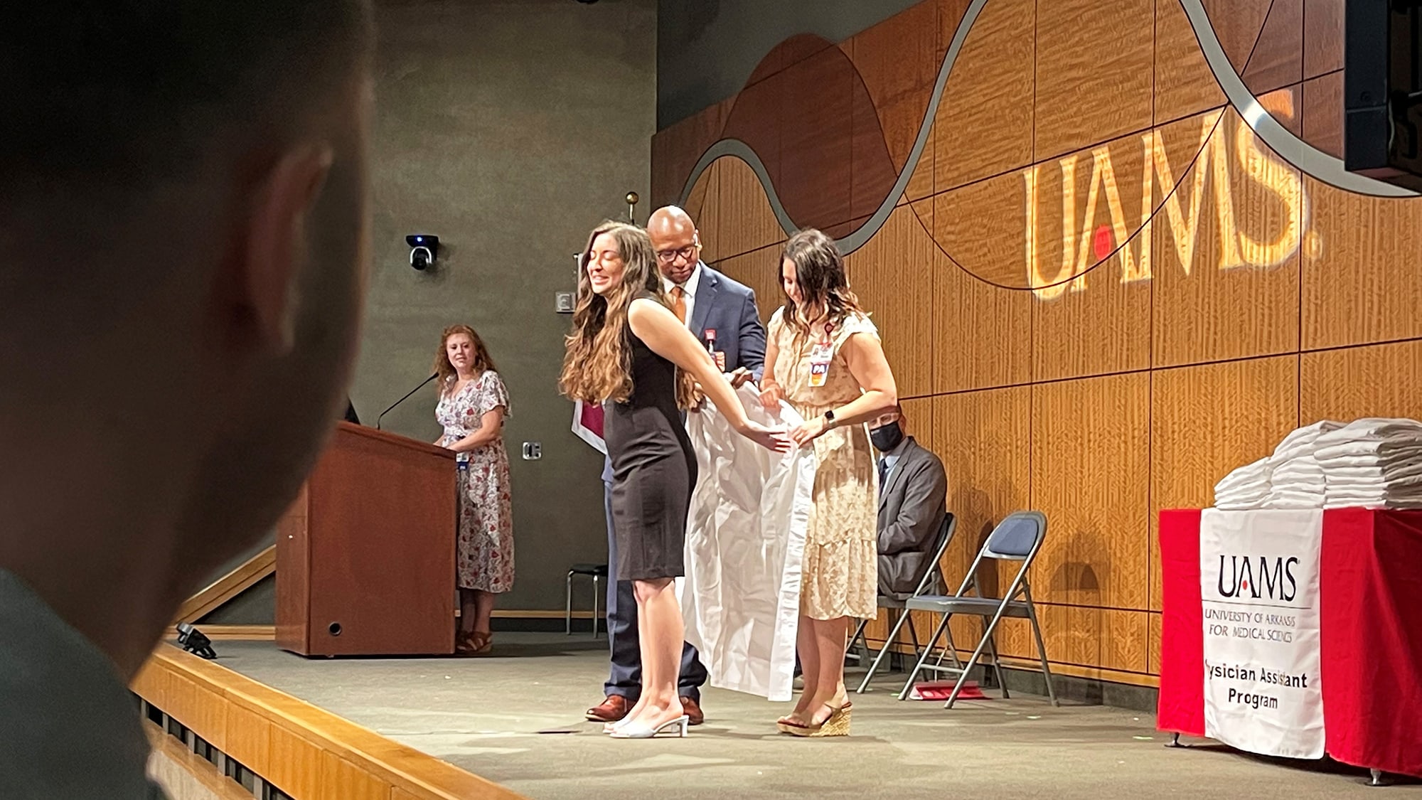 Joseph Frescura, left, waits to step onto the stage and watches as Edward Williams and Hillary Mayberry help Taryn Frazier put on her long, white coat as part of the Valediction Ceremony for the Physician Assistant Program's Class of 2022.
