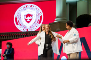 Faculty members help a student put on her white coat during the ceremony.