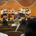 Eleven students in white coats sing on stage as their classmates watch from audience in auditorium.