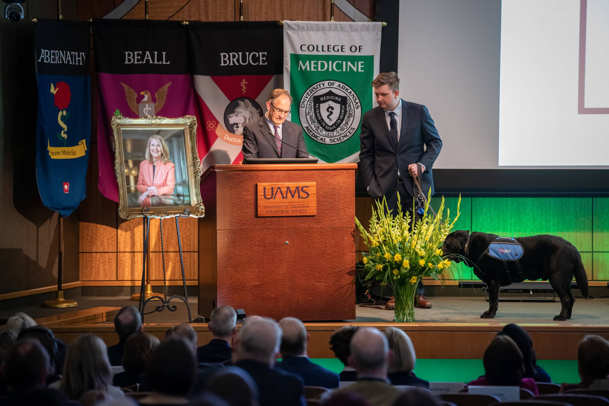 Andrew Morris, Ph.D., addresses the crowd, flanked by his late wife's portrait and son Edward, as well as the family dogs Saleh and Carmine.