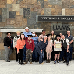 Parents and children in a cancer support group pose for a photo in front of a cancer center sign