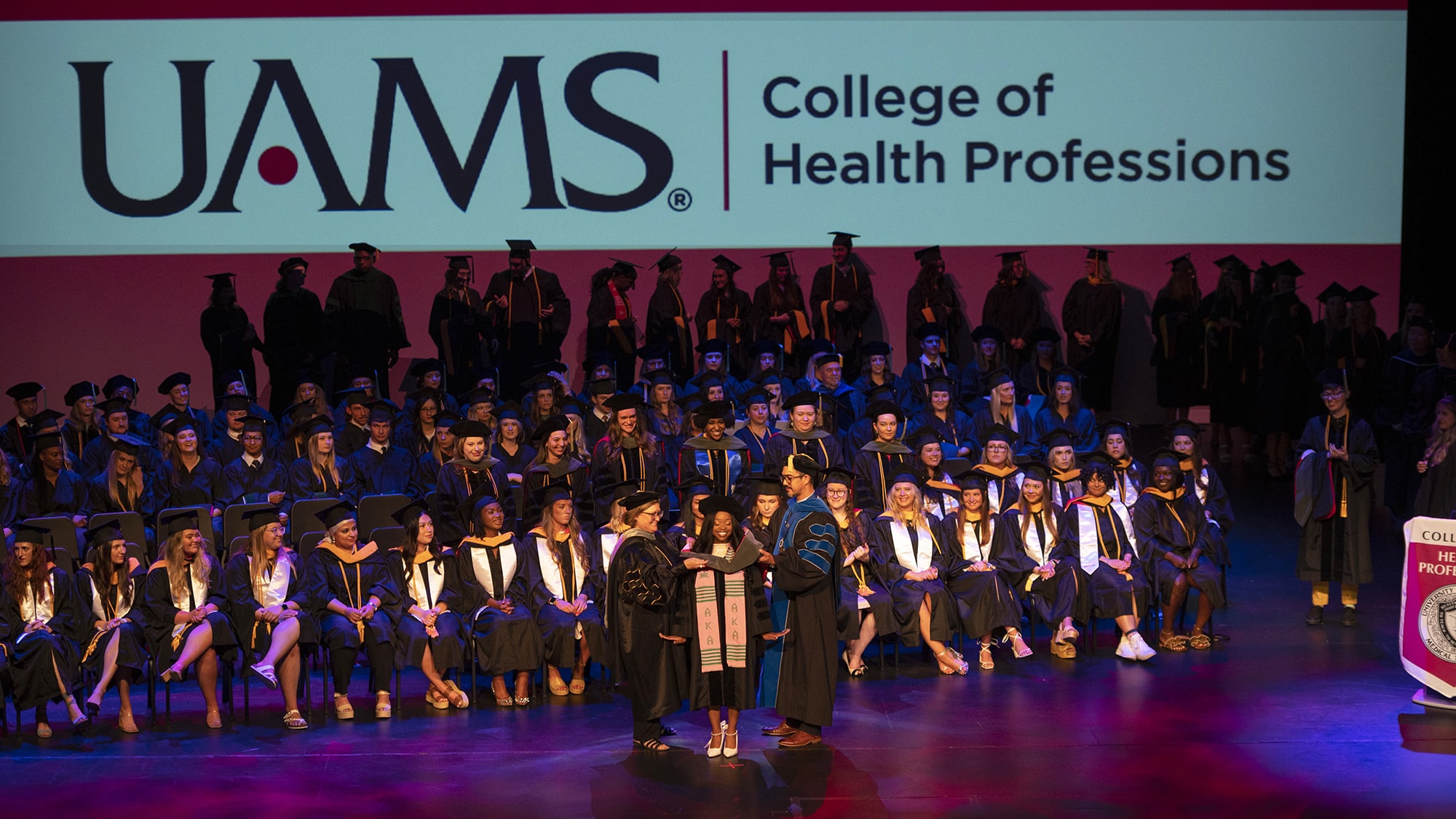 A student stands center stage May 26 in the CHARTS Theater as two members of the College of Health Professions faculty place her academic hood on her shoulders.