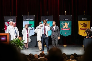 Richard Smith, M.D., watches from the left as students walk in front of flags representing the College of Medicine's academic houses to be helped into their white coats by family members and advisers.