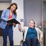 Front view of two businesswomen on their way to a meeting, conversing. One of them is a person using a self-propelled wheelchair; the other is holding a note pad. They are in an office reception area.