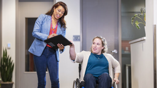 Front view of two businesswomen on their way to a meeting, conversing. One of them is a person using a self-propelled wheelchair; the other is holding a note pad. They are in an office reception area.