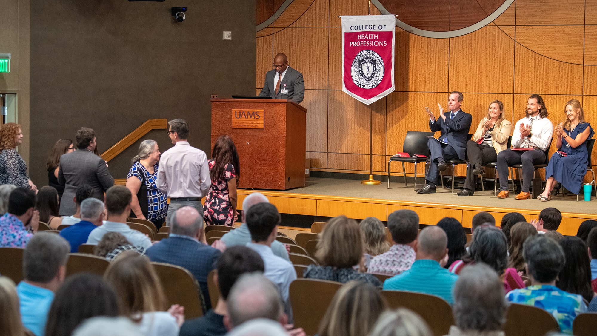 Physician Assistant Studies faculty members stand and receive applause at the valediction ceremony for the Class of 2023. Edward Williams stands and the lectern, left, as Chancellor Cam Patterson, Dean Susan Long, Ryan Toliver and Tiffany Huitt join in the applause.