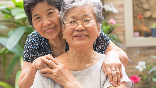 An older woman leans forward and drapes her arms around the shoulders of a woman in front of her.