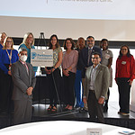 photo of 14 people standing on stage with plaque: Rohit Dhall, M.D., chair of the Department of Neurology (in mask), accepts the plaque from Elizabeth Guerrero of the Parkinson's Foundation (directly to the right of the plaque), while surrounded by team members including Aditya Vikram Boddu, M.D. (bottom right), who co-chairs the UAMS Comprehensive Care Center with Dhall, and Tuhin Virmani, M.D., Ph.D. (standing behind Boddu to the left), division chief of neurodegenerative disorders.
