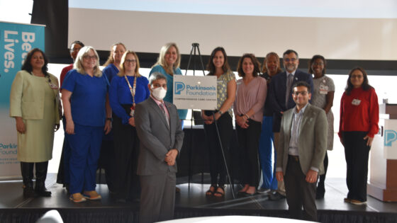 photo of 14 people standing on stage with plaque: Rohit Dhall, M.D., chair of the Department of Neurology (in mask), accepts the plaque from Elizabeth Guerrero of the Parkinson's Foundation (directly to the right of the plaque), while surrounded by team members including Aditya Vikram Boddu, M.D. (bottom right), who co-chairs the UAMS Comprehensive Care Center with Dhall, and Tuhin Virmani, M.D., Ph.D. (standing behind Boddu to the left), division chief of neurodegenerative disorders.
