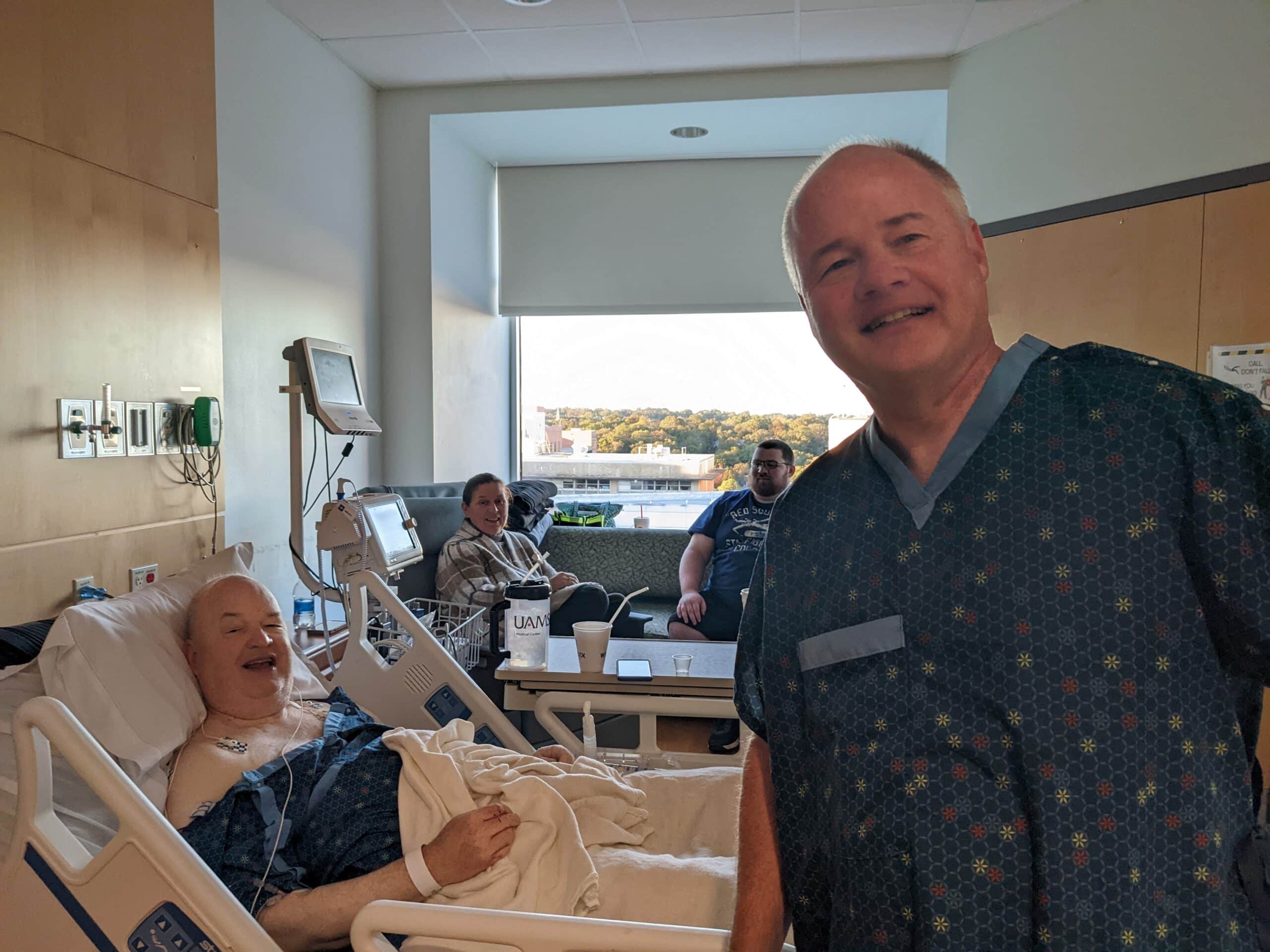 Steve Arhur, standing, in hospital gown, visits with John Arthur, M.D., in a patient bed, after the surgery in which Steve donate a kidney to John.