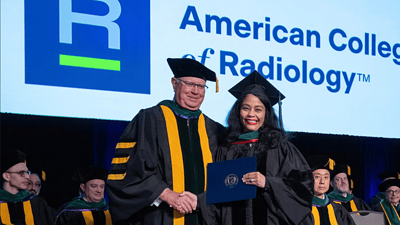 A physician in a cap and gown receives a diploma from a faculty member during the American College of Radiology ceremony, with several people in academic attire seated in the background.