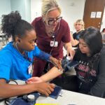 A College of Nursing student wears a blood pressure cuff as another student and an instructor listen through a stethoscope during a classroom exercise.