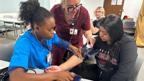 A College of Nursing student wears a blood pressure cuff as another student and an instructor listen through a stethoscope during a classroom exercise.