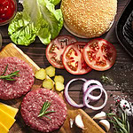 Top view of a rustic wood table filled with ingredients for cooking a delicious homemade cheeseburger. An iron grill with two cooked ground beef burgers is placed at the top-right of the frame and at the opposite side is a cutting board with raw ground beef burgers while the other ingredients are scattered on the table.