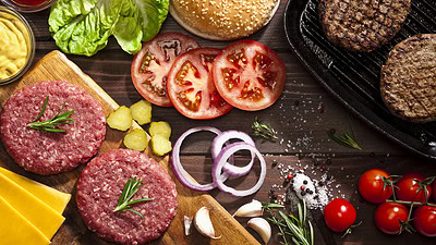 Top view of a rustic wood table filled with ingredients for cooking a delicious homemade cheeseburger. An iron grill with two cooked ground beef burgers is placed at the top-right of the frame and at the opposite side is a cutting board with raw ground beef burgers while the other ingredients are scattered on the table.