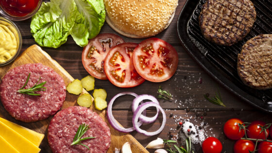 Top view of a rustic wood table filled with ingredients for cooking a delicious homemade cheeseburger. An iron grill with two cooked ground beef burgers is placed at the top-right of the frame and at the opposite side is a cutting board with raw ground beef burgers while the other ingredients are scattered on the table.