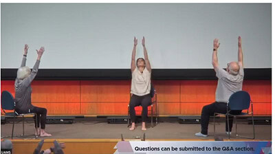 Jessica Holton, occupational therapist, leads two Parkinson's patients in a chair yoga demonstration on the stage as the audience participates from their seats.