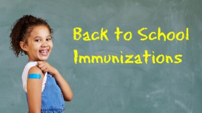 A young child shows off her bandaid while standing in front of the chalkboard that says "Back to School Immunizations"