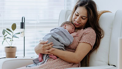 Image of a new mother sitting in a big soft chair holding her newborn wrapped in a blanket.