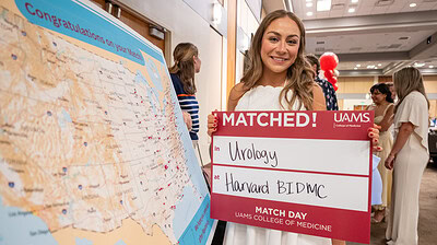Haya Safar stands beside a map showing where all the UAMS seniors are going, holding a sign announcing that she matched at Beth Israel Deaconess Medical Center at Harvard in Boston.