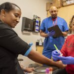 Photo shows three people in an exam room with one preparing a needle for injection and another holding a clipboard.