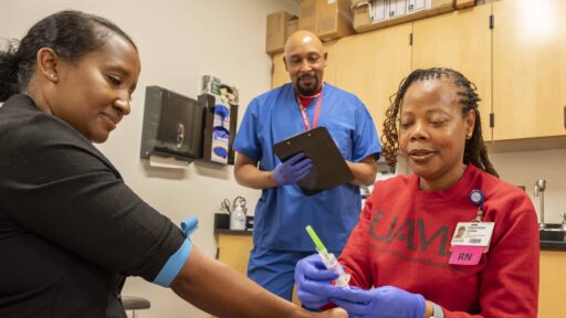 Photo shows three people in an exam room with one preparing a needle for injection and another holding a clipboard.