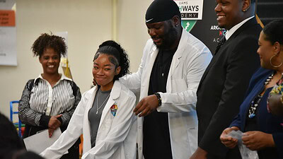 Davonte Hokes, a Hall High and UAMS alumnus and lead pharmacist for Kroger Health, helps a student don her white coat during a ceremony to honor those who took part in the UAMS career development program.