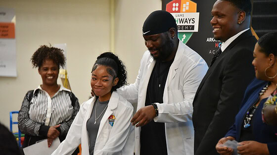 Davonte Hokes, a Hall High and UAMS alumnus and lead pharmacist for Kroger Health, helps a student don her white coat during a ceremony to honor those who took part in the UAMS career development program.