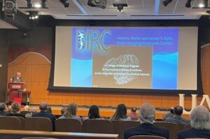 Lee Archer at the podium in front of audience with large image of documents describing neurology research