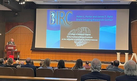Lee Archer at the podium in front of audience with large image of documents describing neurology research