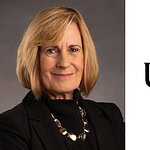 A woman with blonde hair wearing a black blazer and necklace poses against a gray background next to the UAMS logo on a white background.