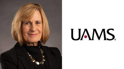 A woman with blonde hair wearing a black blazer and necklace poses against a gray background next to the UAMS logo on a white background.