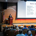 image of Dacre Stoker , next to a big screen with the words 'Bram's vampire traits,' speaking before an audience packed into the auditorium.