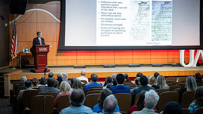 image of Dacre Stoker , next to a big screen with the words 'Bram's vampire traits,' speaking before an audience packed into the auditorium.