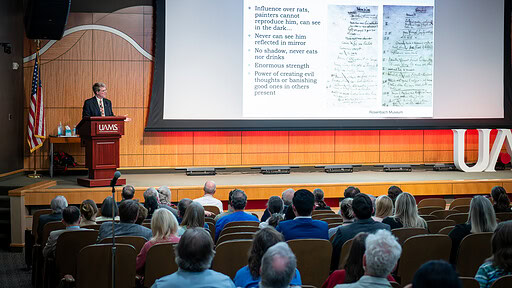 image of Dacre Stoker , next to a big screen with the words 'Bram's vampire traits,' speaking before an audience packed into the auditorium.