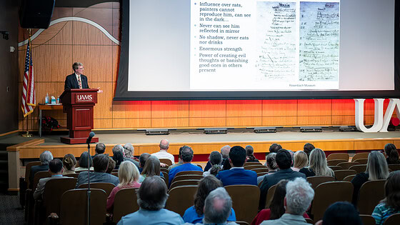 image of Dacre Stoker , next to a big screen with the words 'Bram's vampire traits,' speaking before an audience packed into the auditorium.