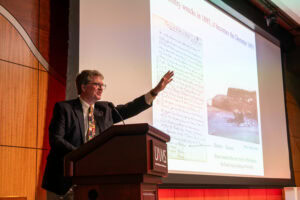 Dacre Stoker with his arm raised at the lectern, next to images of Bram Stoker's handwritten notes.