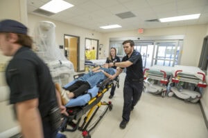 Medics wheel in a woman pretending to be a patient in the emergency room.